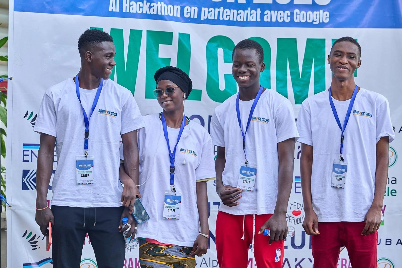 Four young people in ABIHACK 2025 T-shirts with “Staff” badges smile in front of the welcome banner.