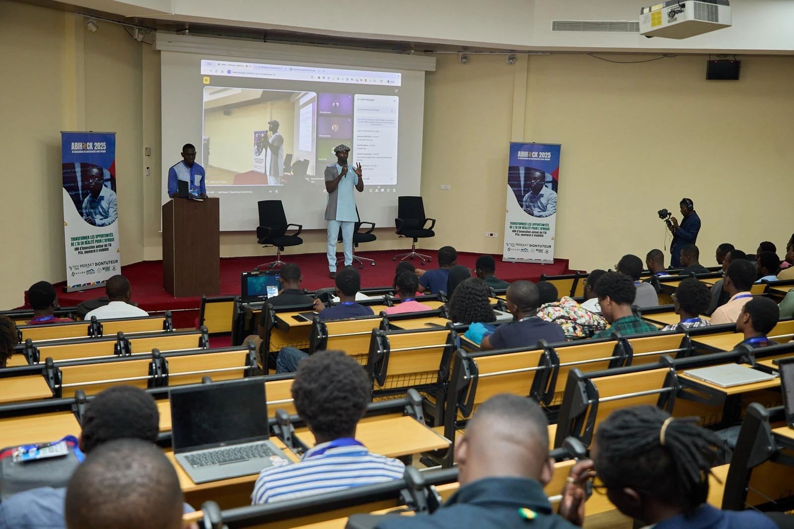 A full lecture hall listens to the speaker on stage, between two ABIHACK 2025 banners.