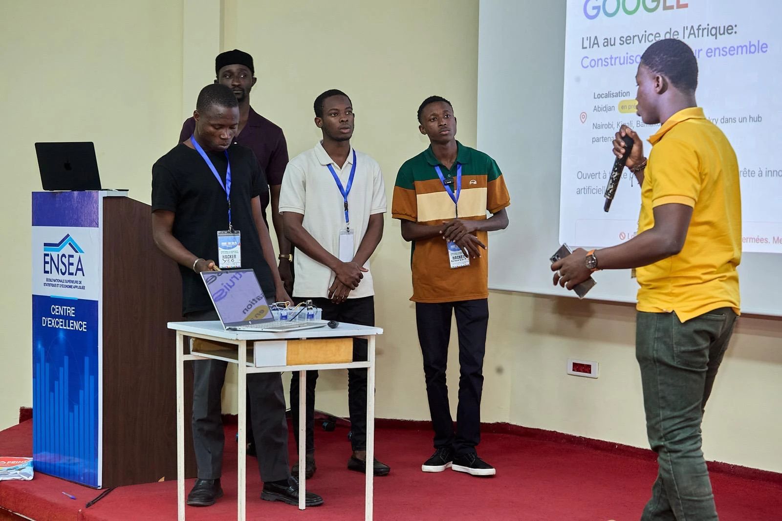 Three young men present their project on stage; a fourth speaks to them into a microphone.