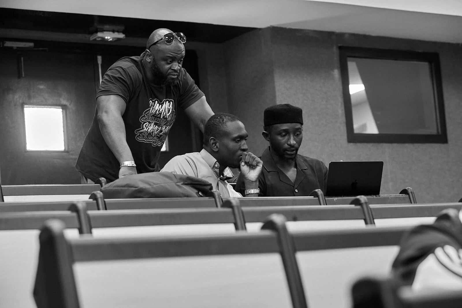 Three men look at a laptop screen together, in a lecture hall.